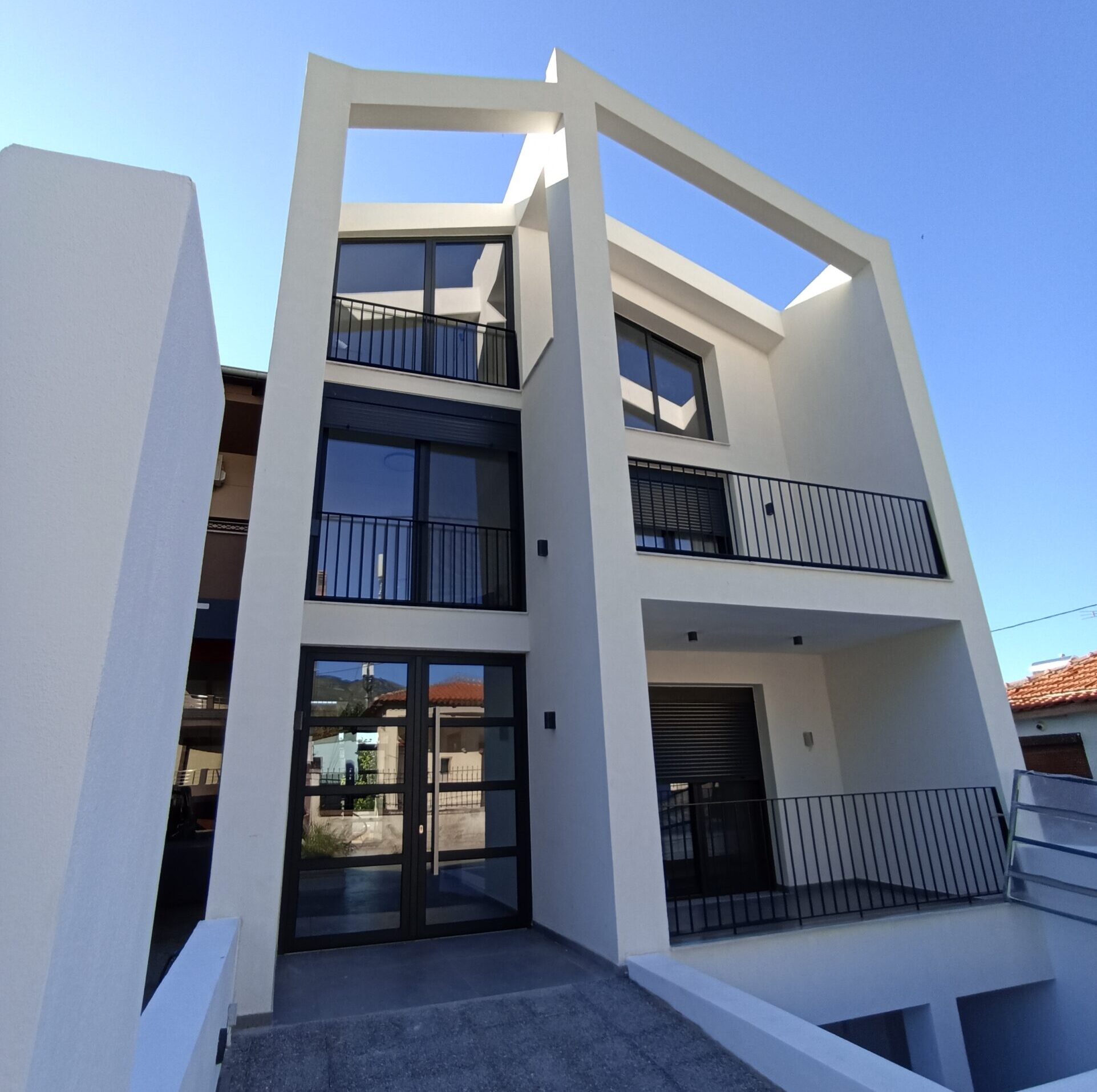 Αρχική όψη απο ένα μοντέρνο λευκό τριώροφο σπίτι με μεγάλα γυάλινα παράθυρα, μαύρα κάγκελα και ξύλινες πέργκολες.