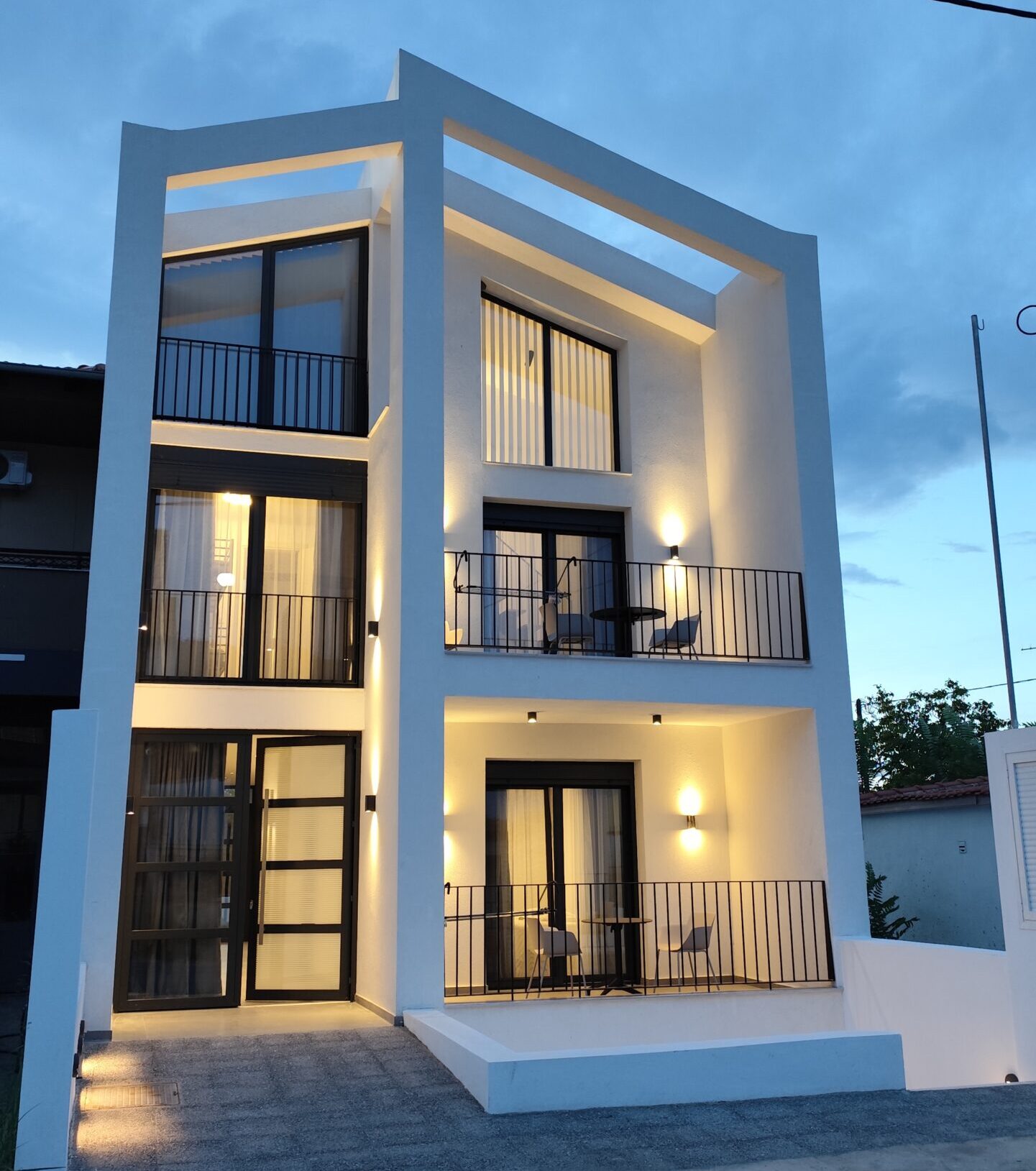 Εναλλακτική όψη απο ένα μοντέρνο λευκό τριώροφο σπίτι με μεγάλα γυάλινα παράθυρα, μαύρα κάγκελα και ξύλινες πέργκολες.